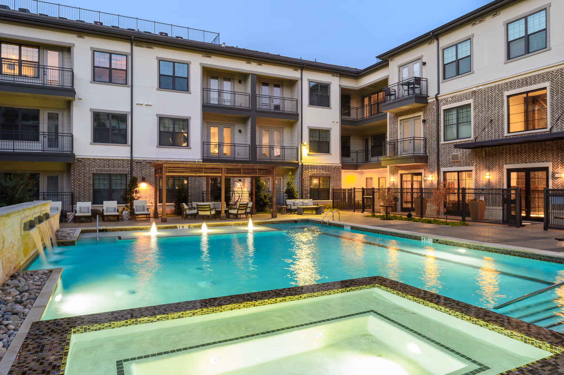Evening view of a modern apartment complex courtyard featuring a large swimming pool with water fountains and a smaller adjacent hot tub. The three-story building surrounds the pool area with balconies and patio seating under a wooden pergola. Warm lights illuminate the scene, creating a welcoming atmosphere.