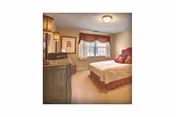 A cozy bedroom in a retirement community featuring a neatly made bed with red and beige bedding, a wooden dresser with a lamp and framed artwork on the wall, a window with curtains allowing natural light into the room, and carpeted flooring.