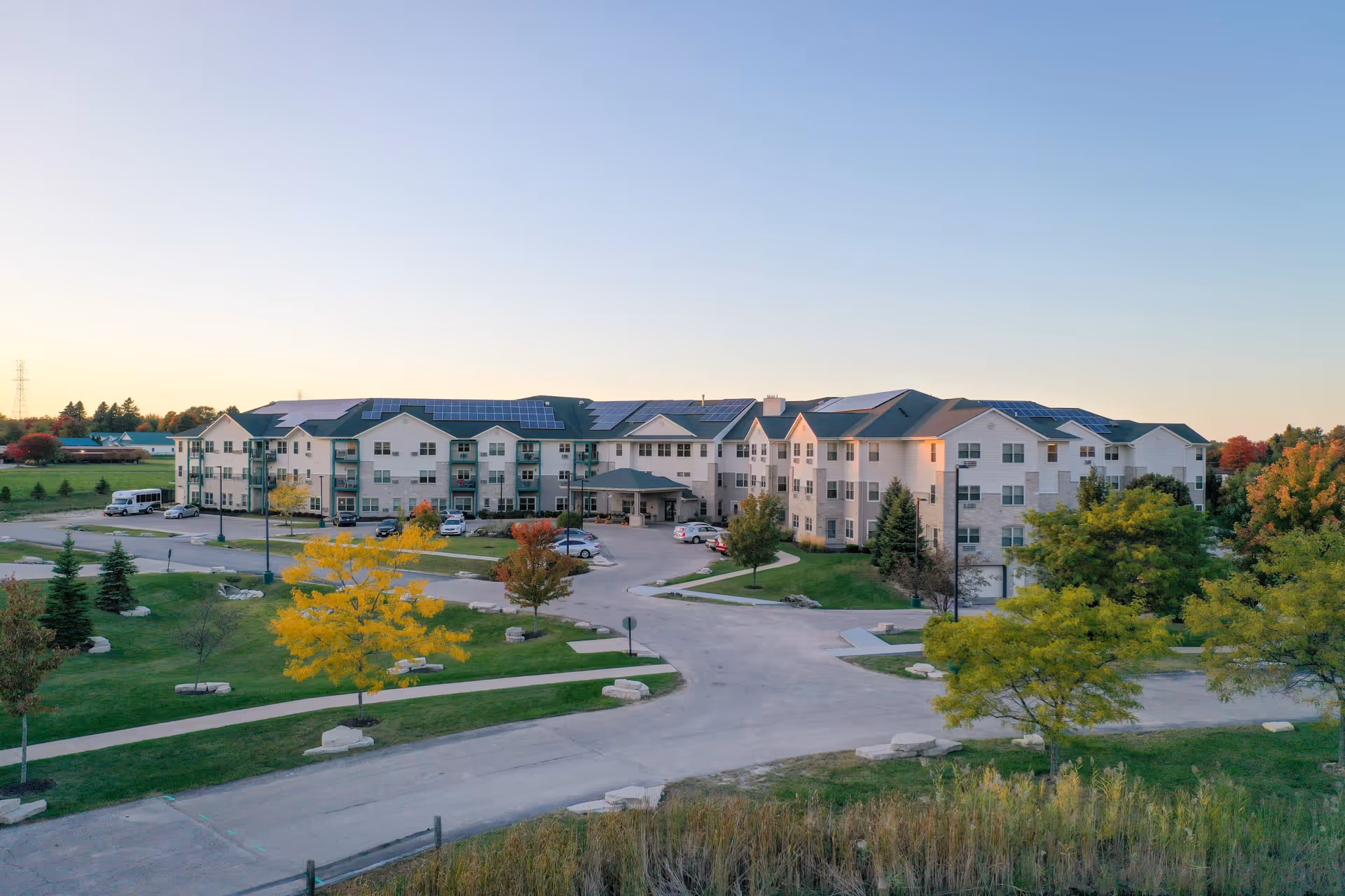 A large, three-story senior living facility building with multiple windows and a covered entrance. The building is surrounded by green lawns, trees with autumn foliage, and paved driveways. Several cars are parked near the entrance, and the sky is clear with a soft gradient from light to deeper blue.