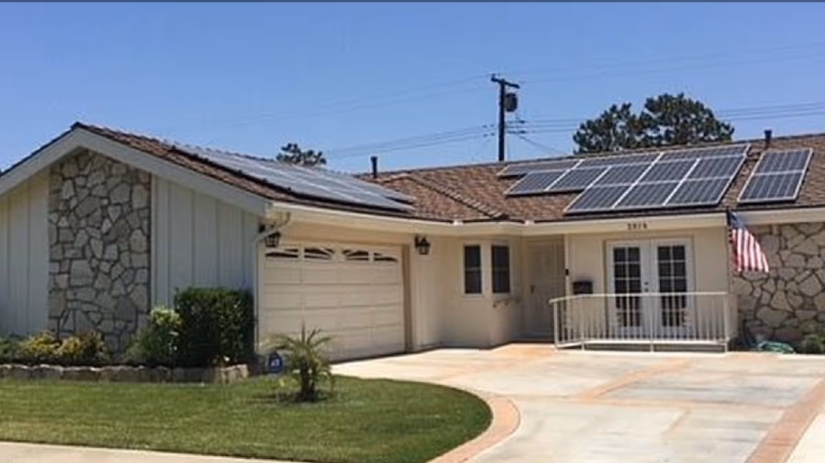 Single-story residential building with a stone and siding exterior, solar panels on the roof, a garage door, a ramp leading to a double-door entrance, and an American flag mounted near the entrance. The front yard has a well-maintained lawn and a small palm plant.