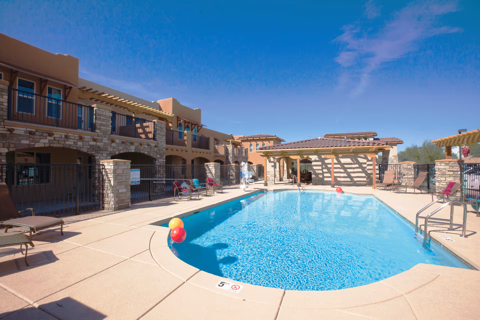 Outdoor swimming pool area at Desert Springs Gracious Retirement Living with clear blue water, surrounded by lounge chairs and a stone and stucco building under a bright blue sky.