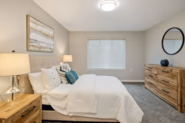 Neatly staged bedroom with a bed dressed in white linens, wooden nightstands and a dresser under a window.