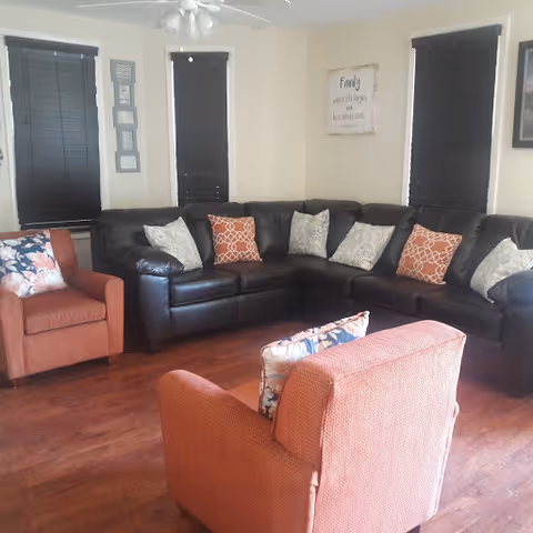 A living room with a large black leather sectional sofa adorned with multiple patterned throw pillows, two orange upholstered armchairs with floral pillows, dark window blinds, wooden flooring, and a ceiling fan. A wall decoration with the word 'Family' is visible on the wall.