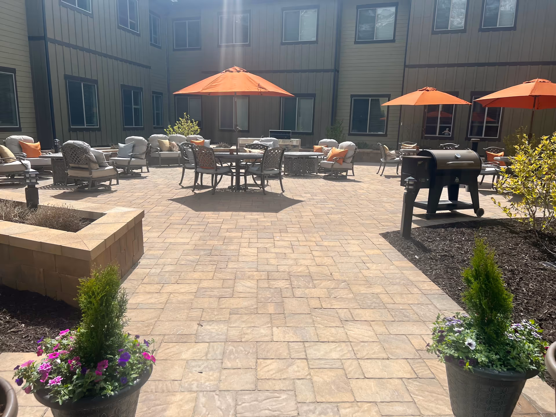 Outdoor patio area at a senior living facility with multiple seating arrangements including cushioned chairs and tables under orange umbrellas. There is a barbecue grill on the right side and potted plants with flowers in the foreground. The building with multiple windows surrounds the patio.
