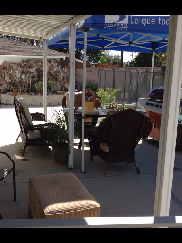Outdoor patio area with a glass-top table surrounded by wicker chairs, potted plants, a barbecue grill, and a blue canopy tent with the DirecTV logo and Spanish text. The area is shaded by a white pergola and there is a rock garden and fence in the background.