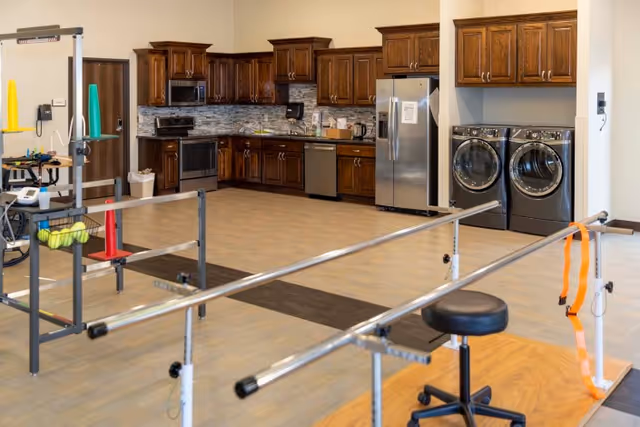 A spacious room featuring a kitchen area with dark wooden cabinets, stainless steel appliances including a refrigerator, oven, microwave, and dishwasher. To the right, there is a laundry area with a front-loading washer and dryer. In the foreground, there are parallel metal handrails used for physical therapy or rehabilitation exercises, along with a black stool and some exercise equipment.