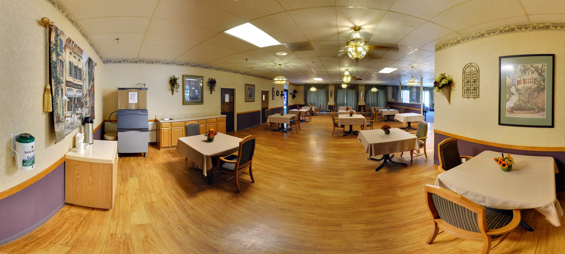 A spacious dining room with multiple tables covered with beige tablecloths and small flower arrangements. The room has wooden flooring, beige walls with purple wainscoting, framed artwork, and decorative wall hangings. Ceiling lights with glass fixtures illuminate the area. There is a water dispenser and a small counter with cups on the left side of the room.