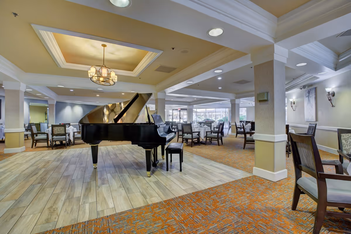 Spacious dining area in Thunderbird Senior Living featuring a black grand piano on a wooden floor section, surrounded by multiple tables and chairs with white tablecloths. The room has a warm color palette with beige walls, recessed lighting, and a decorative ceiling light fixture.