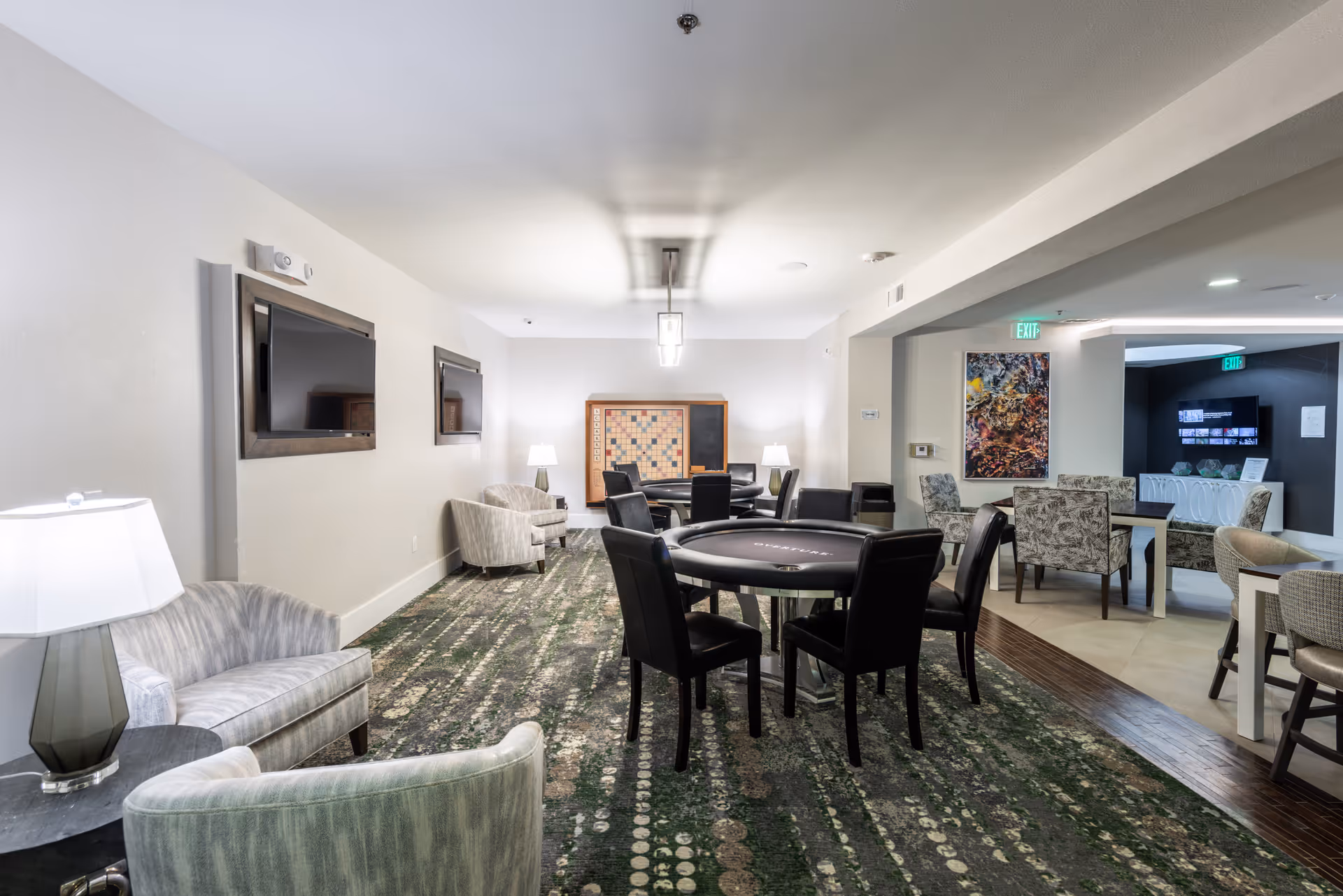 Communal lounge with a central round card table surrounded by chairs, additional seating, wall-mounted TVs, and lamps.