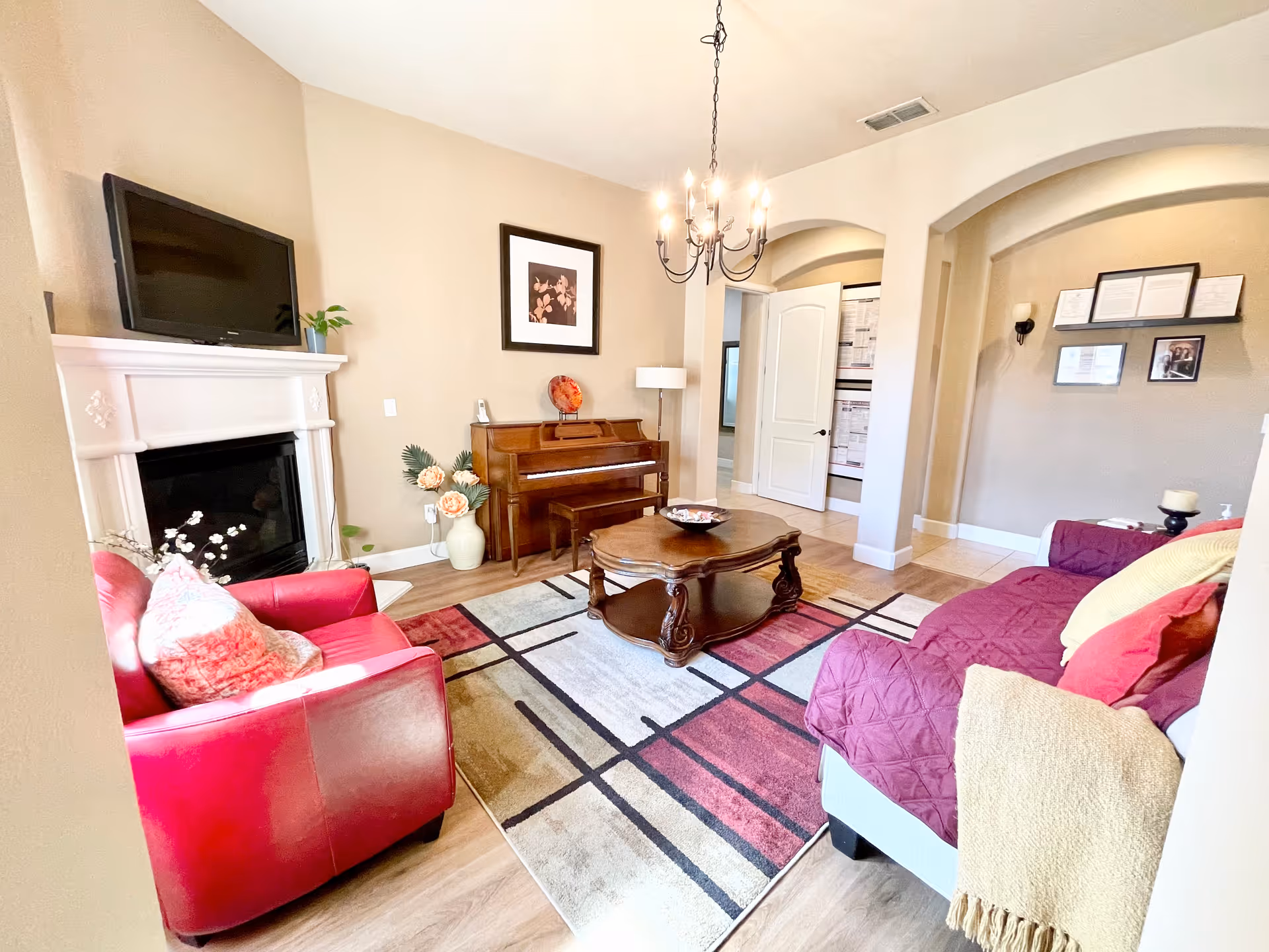 A cozy living room with a red armchair and a red sofa covered with a quilted cover. There is a wooden coffee table on a patterned area rug in the center. A piano is against the wall with a framed picture above it. A white fireplace with a TV on the mantel is on the left side. The room has beige walls, wooden flooring, and a chandelier hanging from the ceiling. There are some decorative plants and framed certificates on the wall in the background.