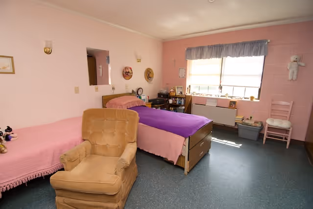 A simple, cozy bedroom in an assisted living facility with two single beds, one covered with a pink blanket and the other with a purple blanket. There is a cushioned armchair in the foreground. The walls are painted light pink, and there is a window with a blue valance letting in natural light. Various personal items and decorations are on the walls and a small shelf near the window.