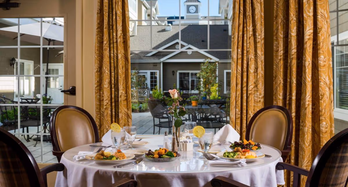 A round dining table set for four with plates of food, glasses of water with lemon, and a vase with a single pink rose. The table is in front of large glass doors with yellow patterned curtains, looking out onto an outdoor patio area with tables, chairs, plants, and a building with a clock tower in the background.