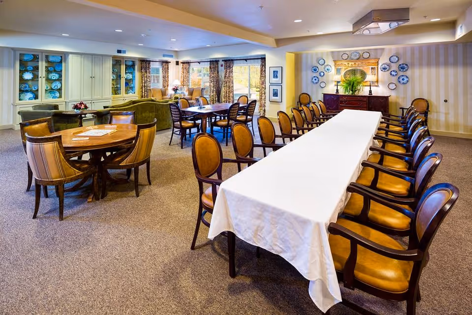 Spacious senior community dining/lounge area with a long table covered in a white tablecloth, multiple round tables and chairs, and display cabinets.