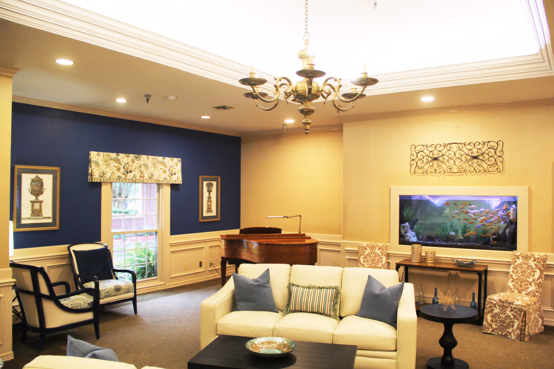 Comfortable senior living common room with sofas, chairs, a piano, chandelier and a built-in aquarium.