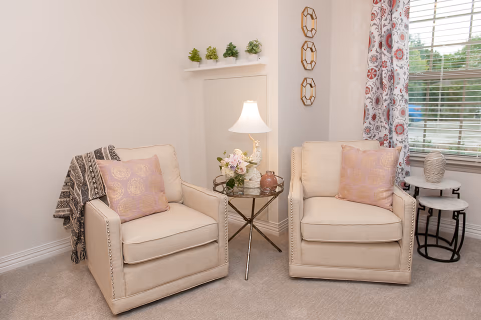 Two beige upholstered armchairs flank a glass side table with a lamp and floral arrangement in a bright living room corner with patterned curtains.