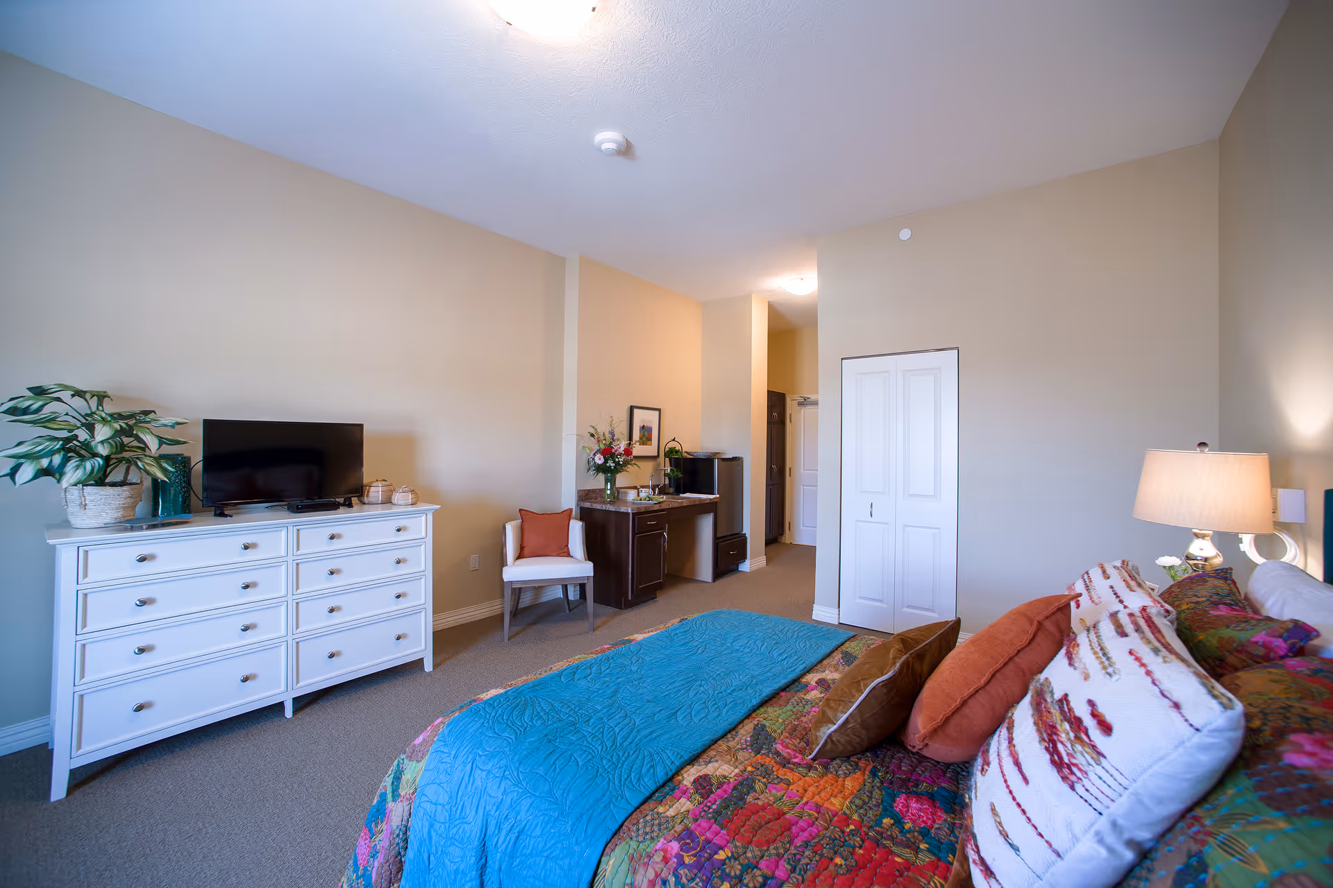 A cozy senior living bedroom featuring a bed with colorful bedding and multiple pillows, a white dresser with a flat-screen TV and a potted plant on top, a white chair with an orange cushion, a small dark wood desk with flowers and a coffee maker, and a closed white closet door. The walls are painted beige and the room is well-lit with a lamp on the bedside table.