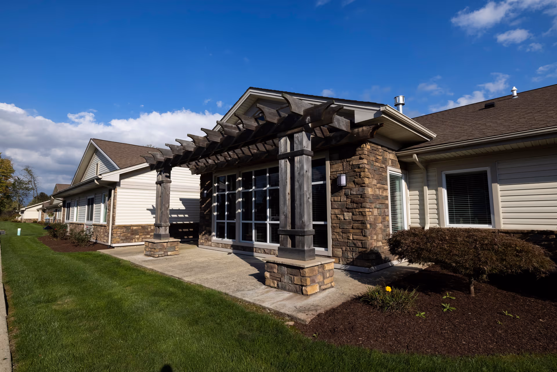Exterior view of Elmwood Senior Care Facility showing a building with beige siding and stone accents. There is a wooden pergola over a concrete patio area, surrounded by well-maintained green grass and small shrubs under a blue sky with some clouds.