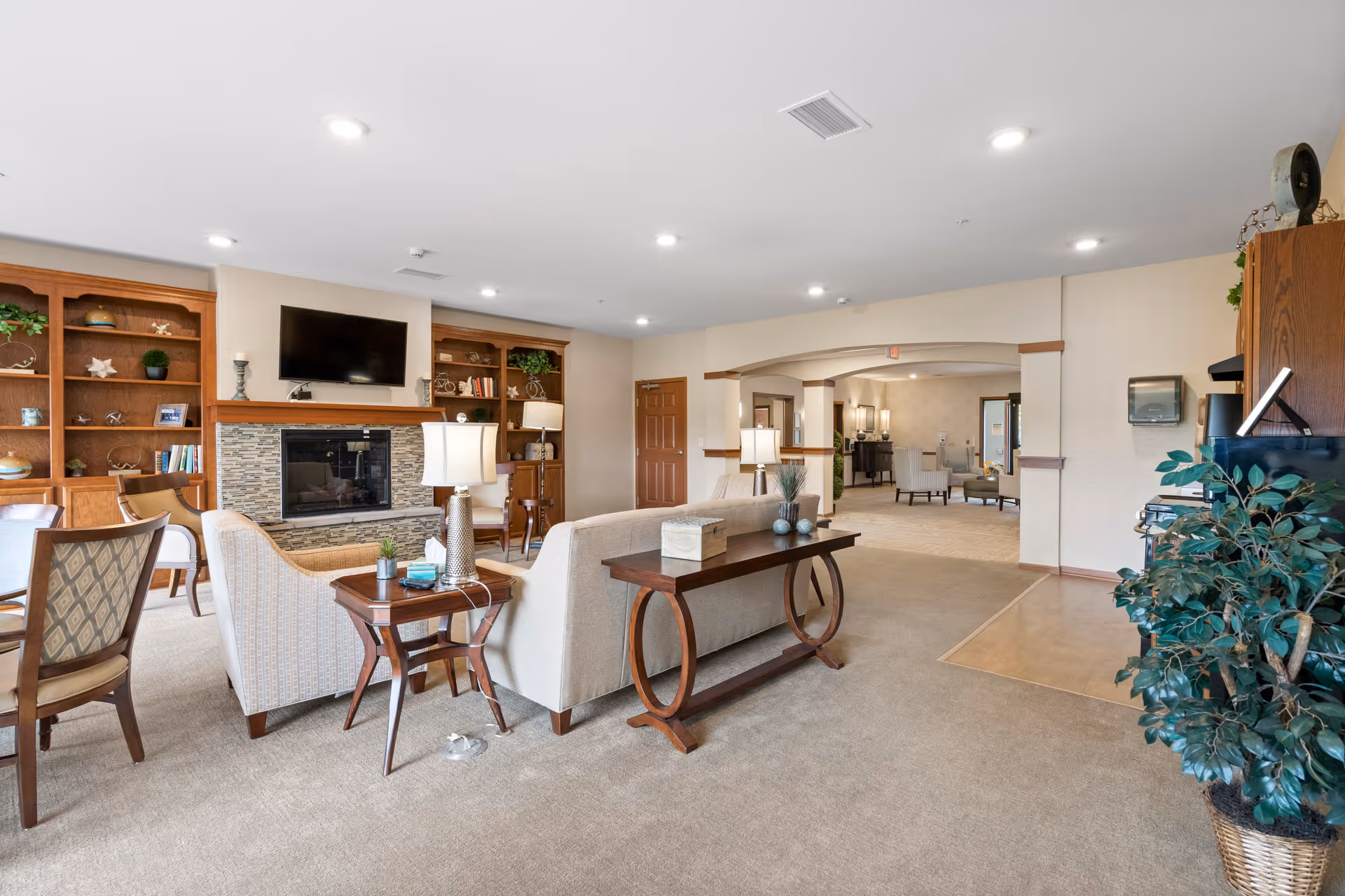 A spacious and well-lit living room area in a senior living facility featuring beige carpeting, a stone fireplace with a mounted flat-screen TV above it, wooden built-in shelves with decorative items, comfortable upholstered chairs and sofas, side tables with lamps, and a large potted plant near the entrance to another room.