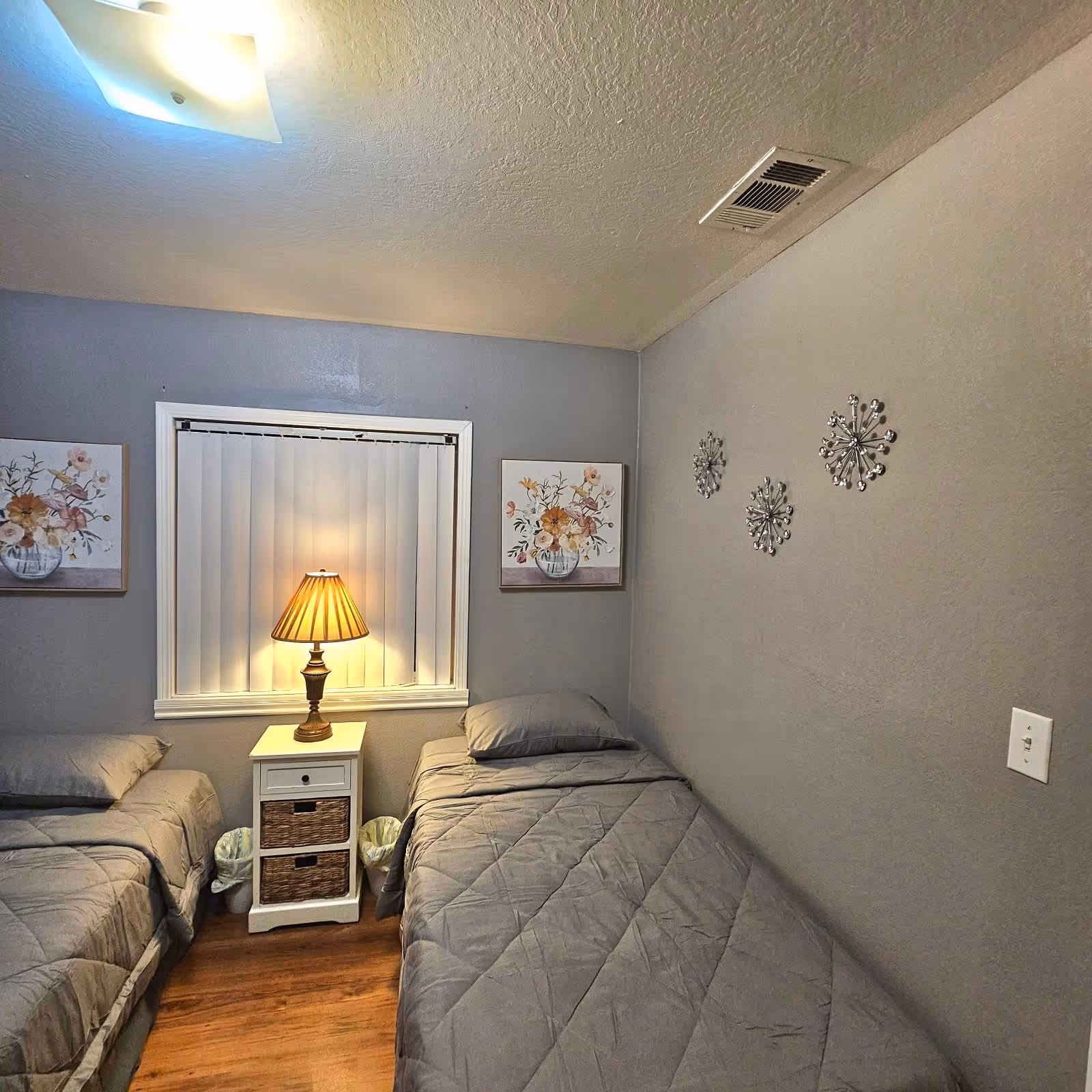 A small bedroom with two single beds covered in gray bedding. Between the beds is a white nightstand with two wicker drawers and a table lamp with a beige shade. The walls are painted gray and decorated with floral paintings and metallic wall art. The floor is wooden, and there is a window with vertical blinds behind the nightstand.