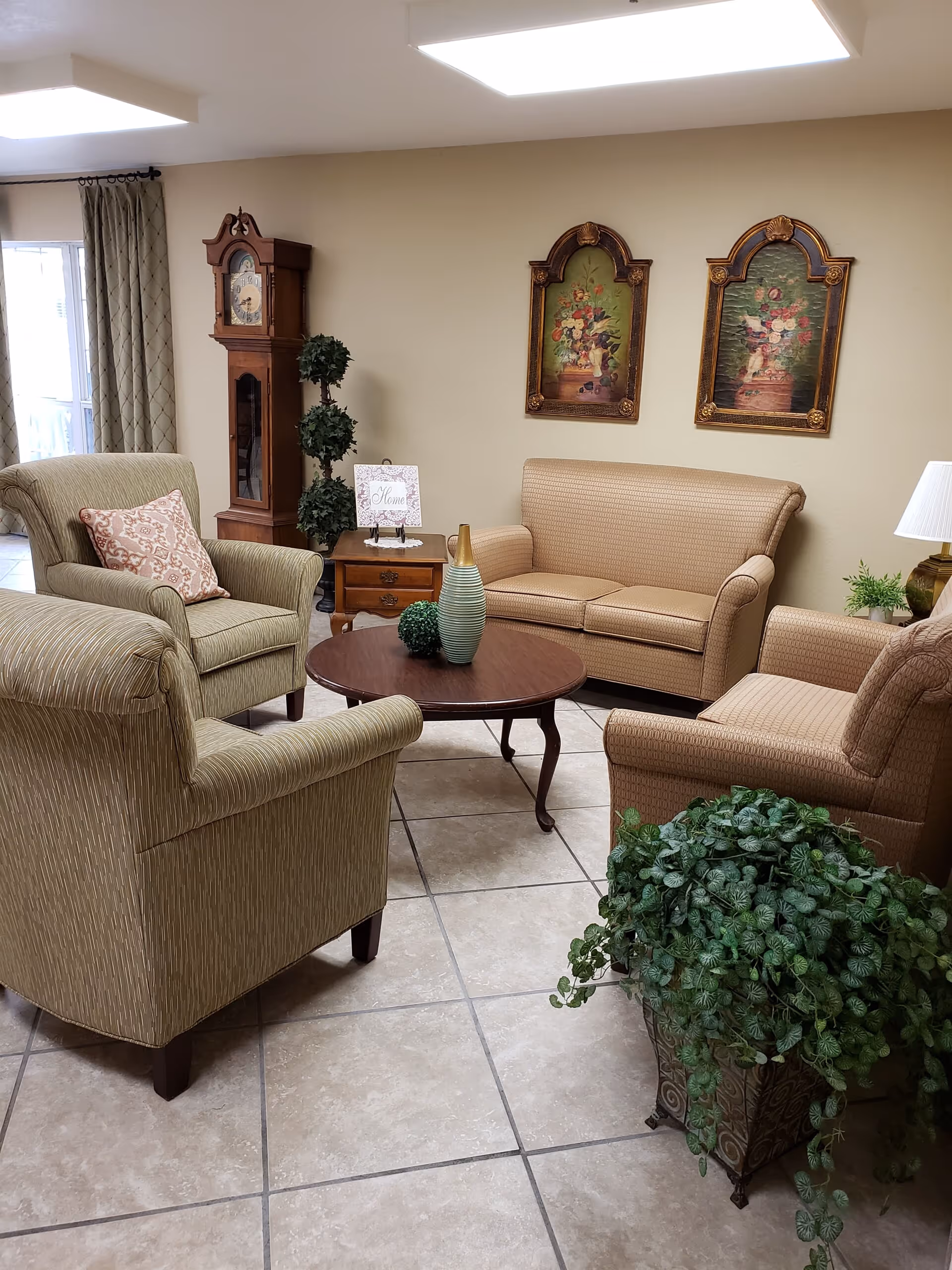 A cozy living room area with two beige armchairs, a beige loveseat, a round wooden coffee table with decorative vases, a wooden grandfather clock, two framed floral paintings on the wall, a small wooden side table with a 'Home' sign, a table lamp, and a large potted green plant on tiled flooring.