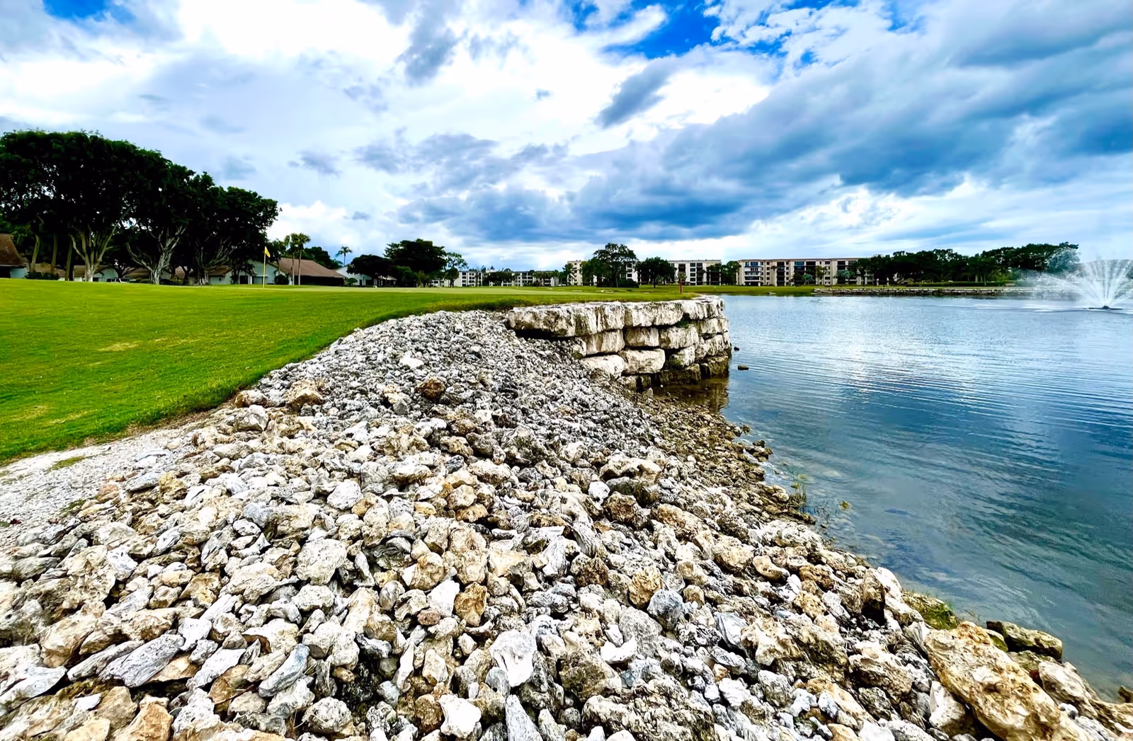 A scenic outdoor view of a rocky shoreline next to a calm lake with a water fountain in the distance. There is a well-maintained grassy area with trees and buildings visible in the background under a partly cloudy sky.
