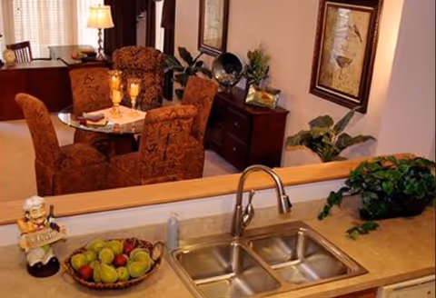 Kitchen sink and countertop with a bowl of fruit and faucet overlooking a dining area with upholstered chairs and a table.