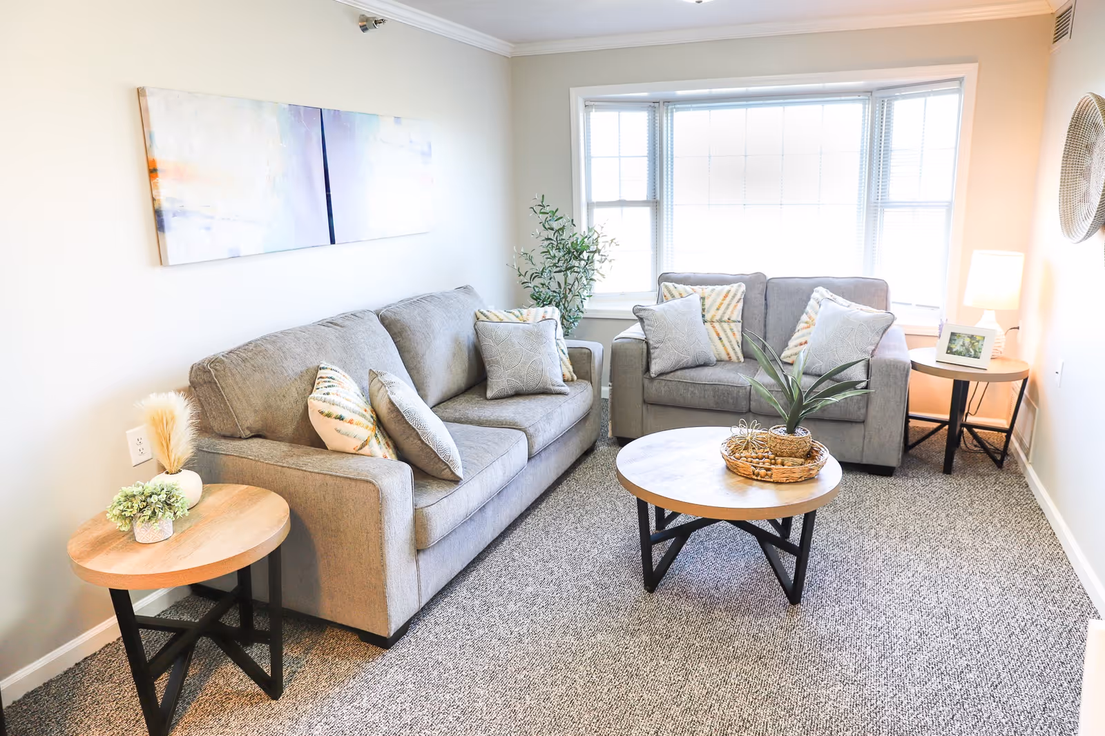 A bright living room with two gray sofas adorned with patterned throw pillows. There are two round wooden side tables, one with a lamp and framed photo, and the other with decorative plants. A round wooden coffee table with a plant centerpiece sits in the middle on a carpeted floor. A large window lets in natural light, and a modern abstract painting hangs on the wall.