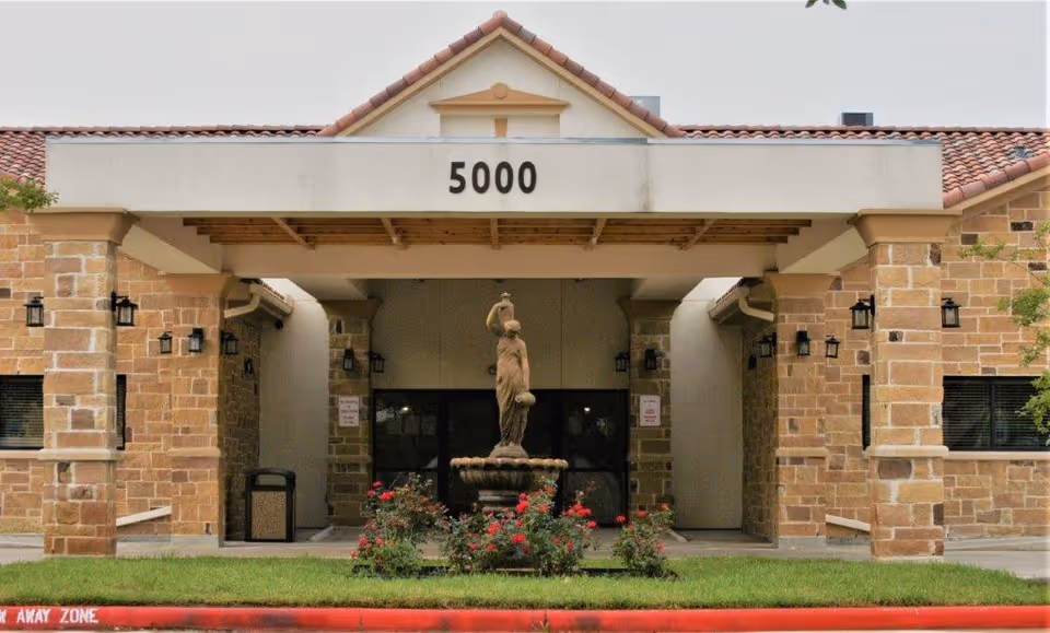Front entrance of a stone-clad building with '5000' above a covered portico and a central fountain statue surrounded by flowers.