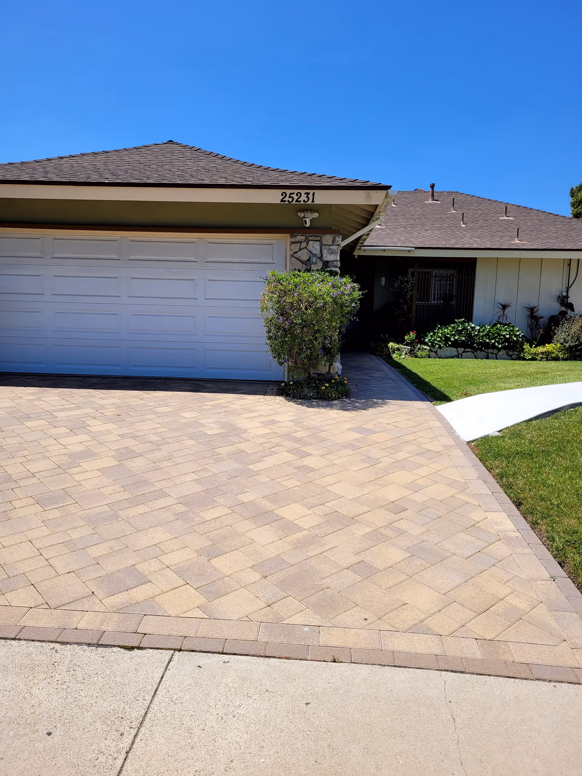 Front exterior view of a single-story house with a double garage door, a paved driveway, a small bush near the garage, and a well-maintained lawn with plants along the side. The house number 25231 is visible above the garage door under a clear blue sky.