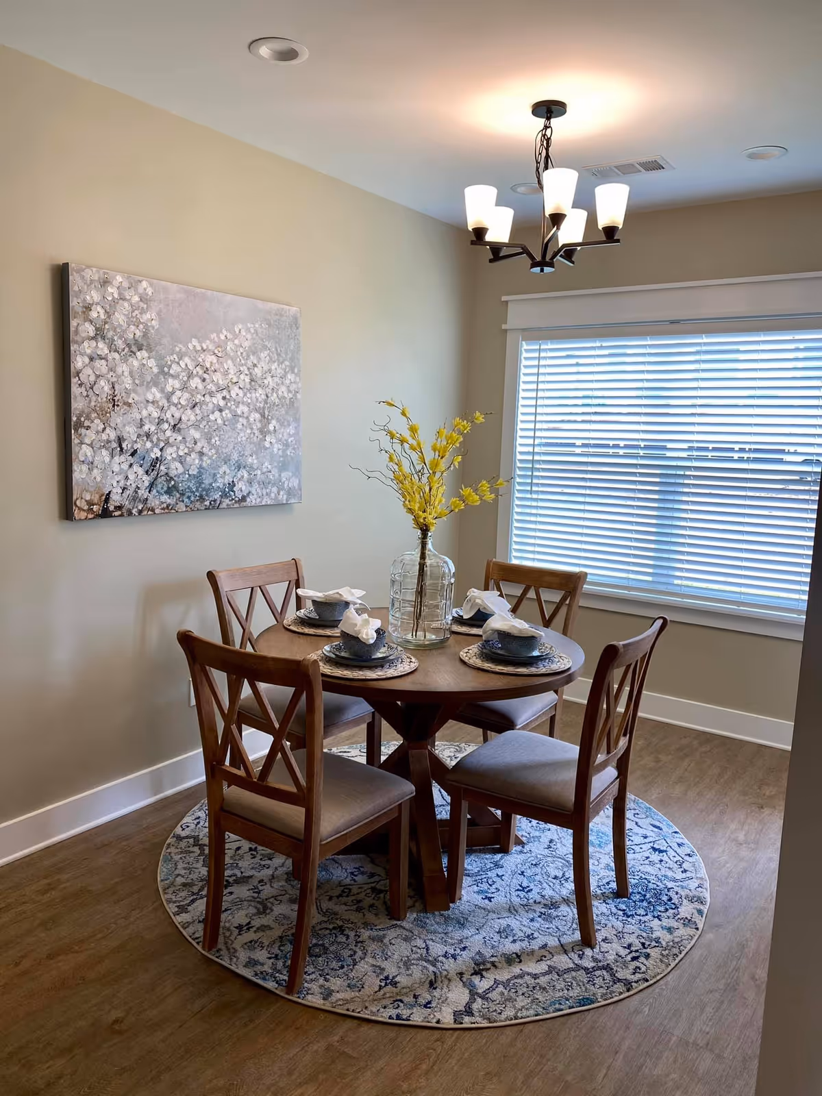 Small dining area with a round wooden table set for four, chairs on a patterned round rug, a vase with yellow flowers, and a chandelier by a window.