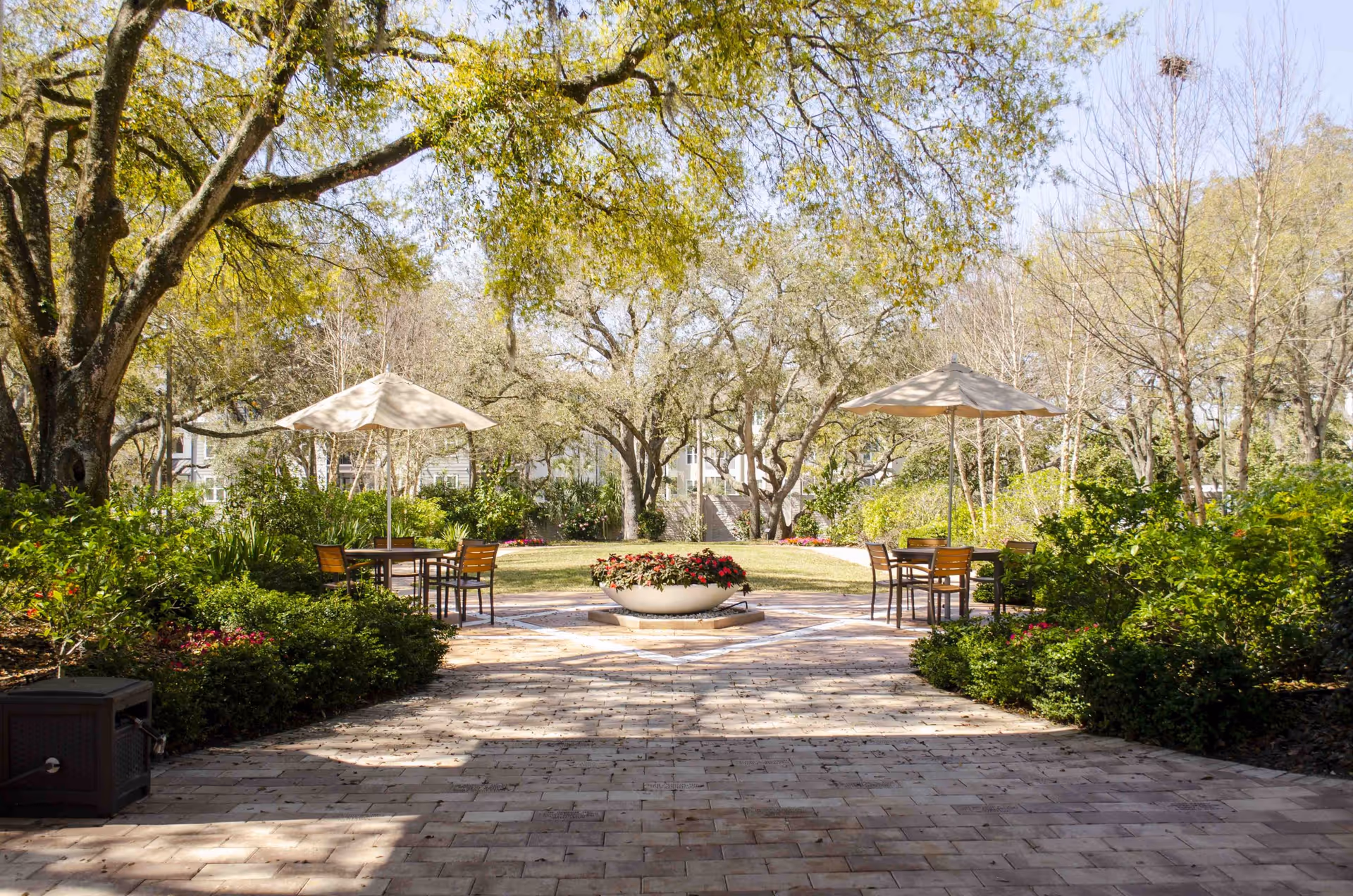 A peaceful outdoor garden area with a paved walkway leading to a circular flower bed filled with red flowers. There are two tables with umbrellas and chairs on either side of the walkway, surrounded by lush green bushes and trees under a clear sky.