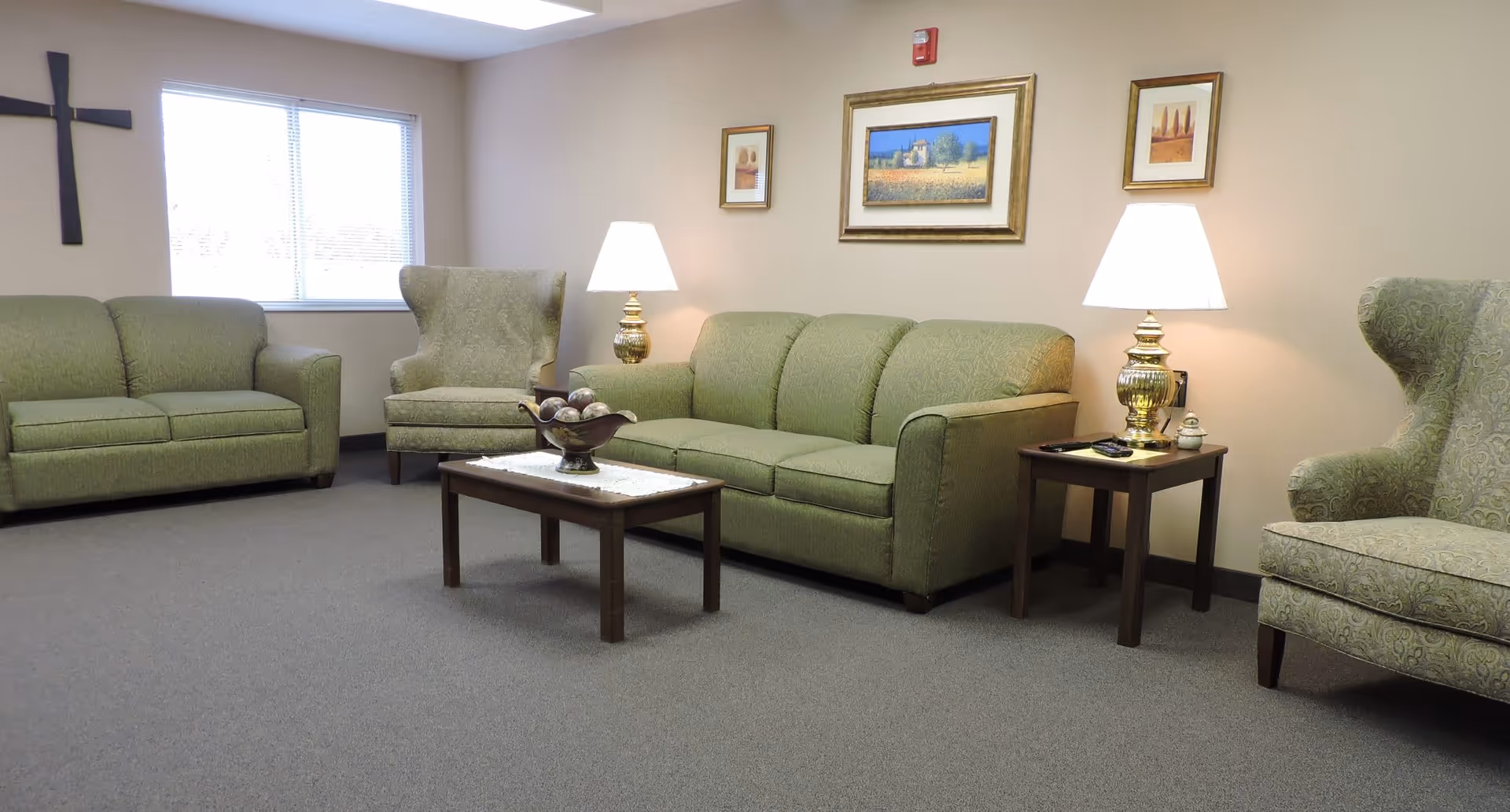 A cozy living room with green upholstered sofas and armchairs, a wooden coffee table with a decorative bowl, two side tables with lamps, framed artwork on the wall, a window with blinds, and a wall-mounted cross.