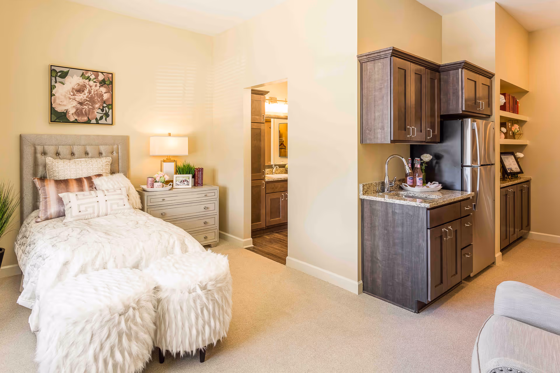 A cozy assisted living room featuring a single bed with multiple pillows and a tufted headboard, a nightstand with a lamp, books, and framed photos, two fluffy white ottomans at the foot of the bed, and a kitchenette with dark wood cabinets, a granite countertop, a sink, and a stainless steel refrigerator. The room has beige walls and carpet, with a floral painting above the bed and an open doorway leading to a bathroom.