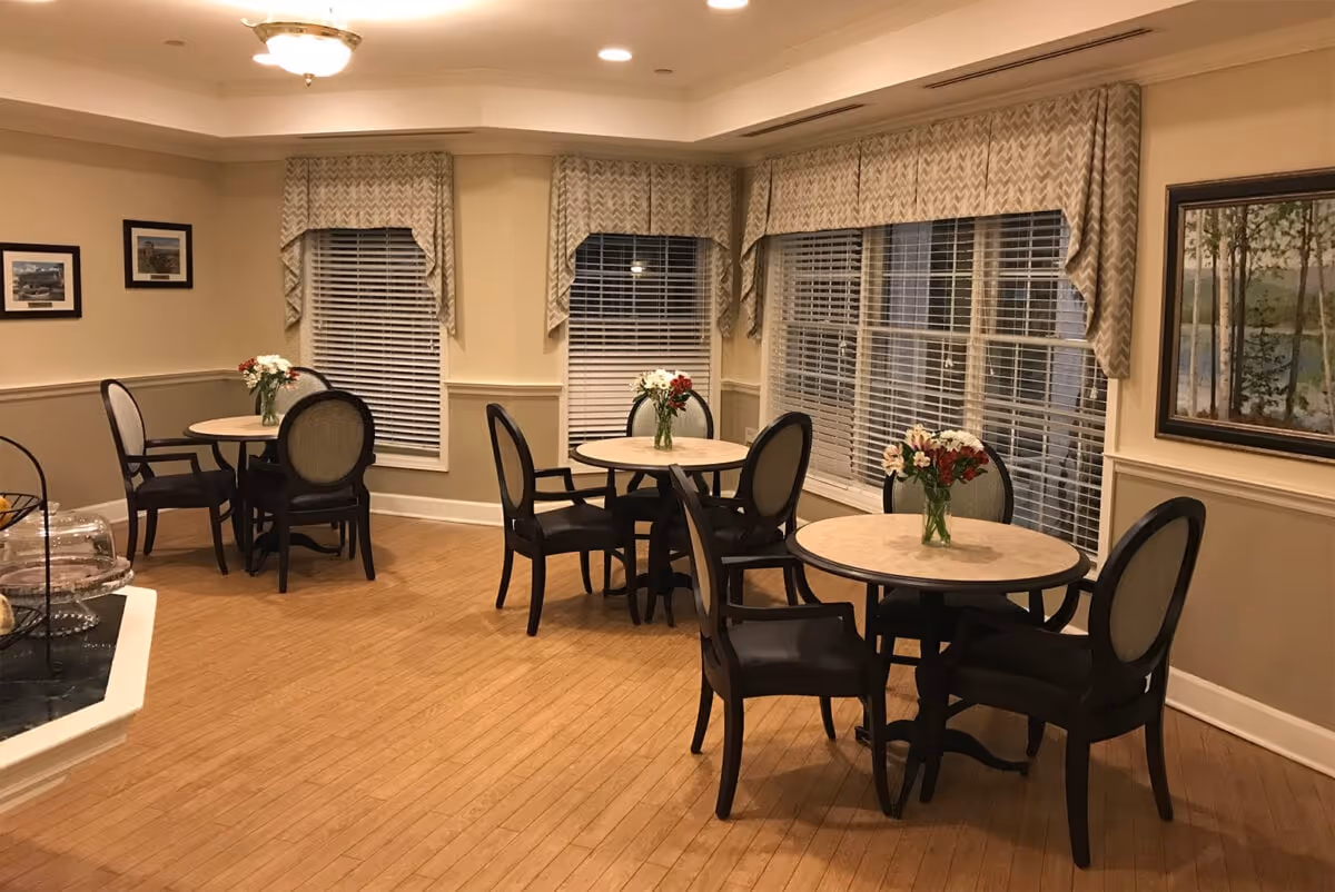 A cozy dining area with three round tables, each surrounded by four chairs. Each table has a small vase with flowers. The room has large windows with blinds and patterned valances, light-colored walls with wainscoting, and framed artwork. The floor is wooden, and the lighting is warm and inviting.