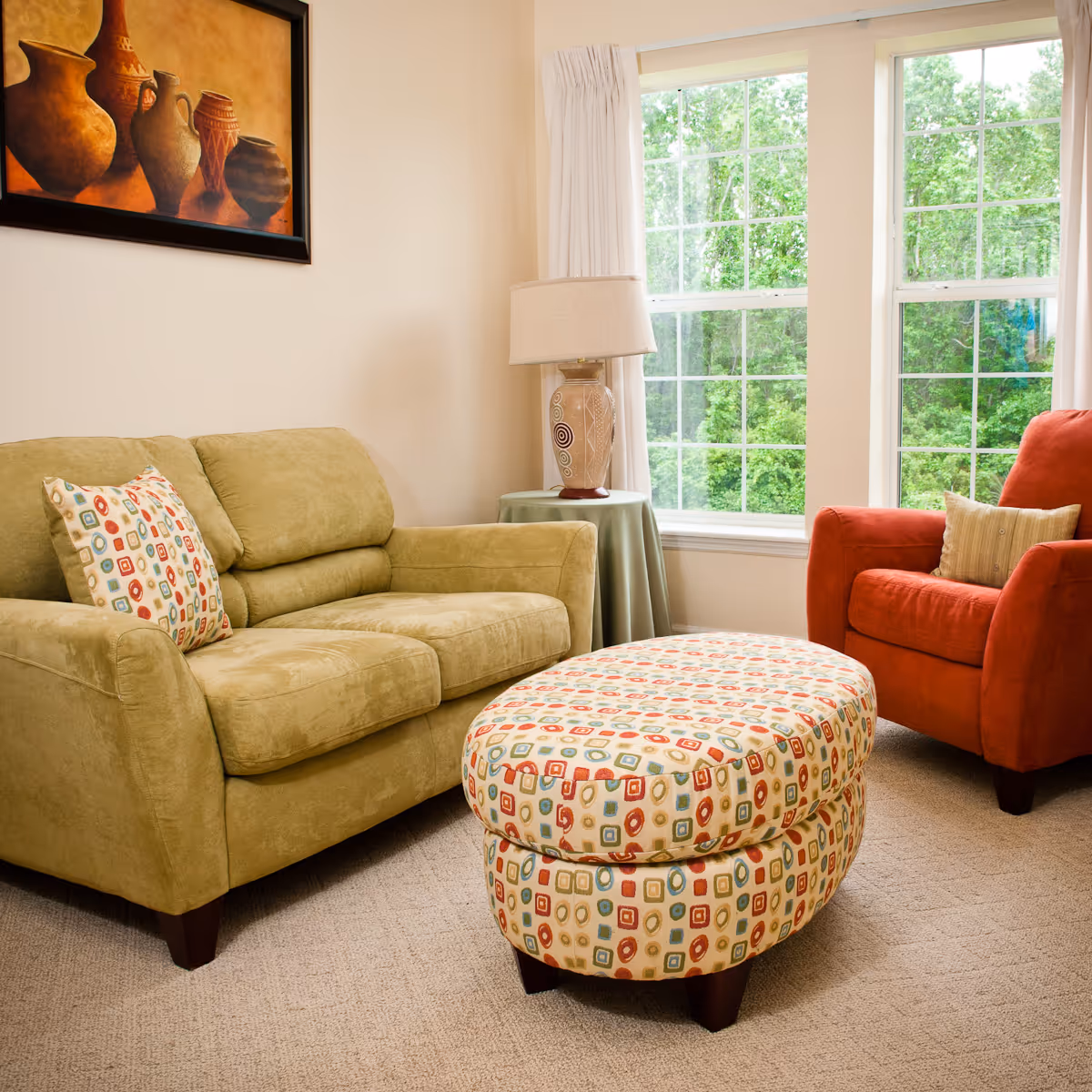 A cozy living room with a green loveseat, a red armchair, and a patterned ottoman in the center. There is a side table with a decorative lamp next to the loveseat, and large windows with white curtains letting in natural light. A framed painting of pottery hangs on the wall above the loveseat.