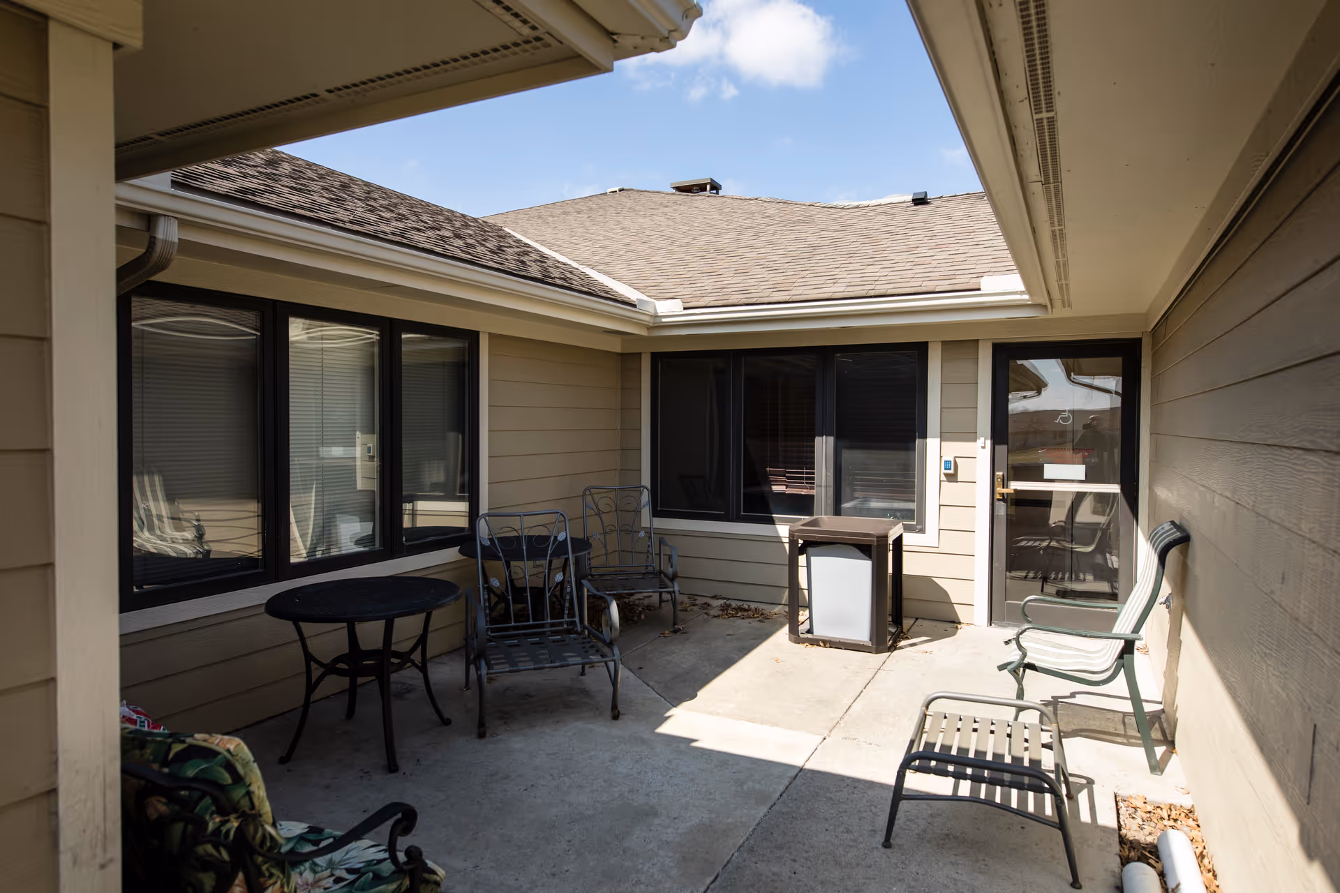 A small outdoor patio area enclosed by beige siding walls and windows. The patio has a concrete floor and contains several metal chairs, a small round table, and a small square table. The sky is clear and blue above the roofline.