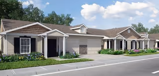 Exterior view of a single-story residential building with beige siding, dark shutters, and a brown shingled roof. The building has multiple entrances, a garage, and landscaped greenery with flowers along the front. The sky is partly cloudy.