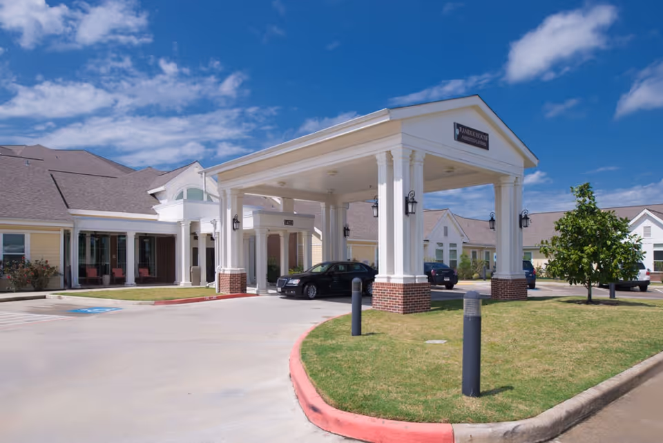 Front entrance of a retirement community with a covered porte-cochere, parked cars, and landscaped lawn under a blue sky.