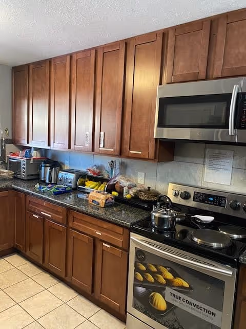 A kitchen with wooden cabinets, a granite countertop, and various kitchen appliances including a microwave, toaster oven, electric kettle, and stove with a pot on it. There are bananas, bread, and other food items on the counter.