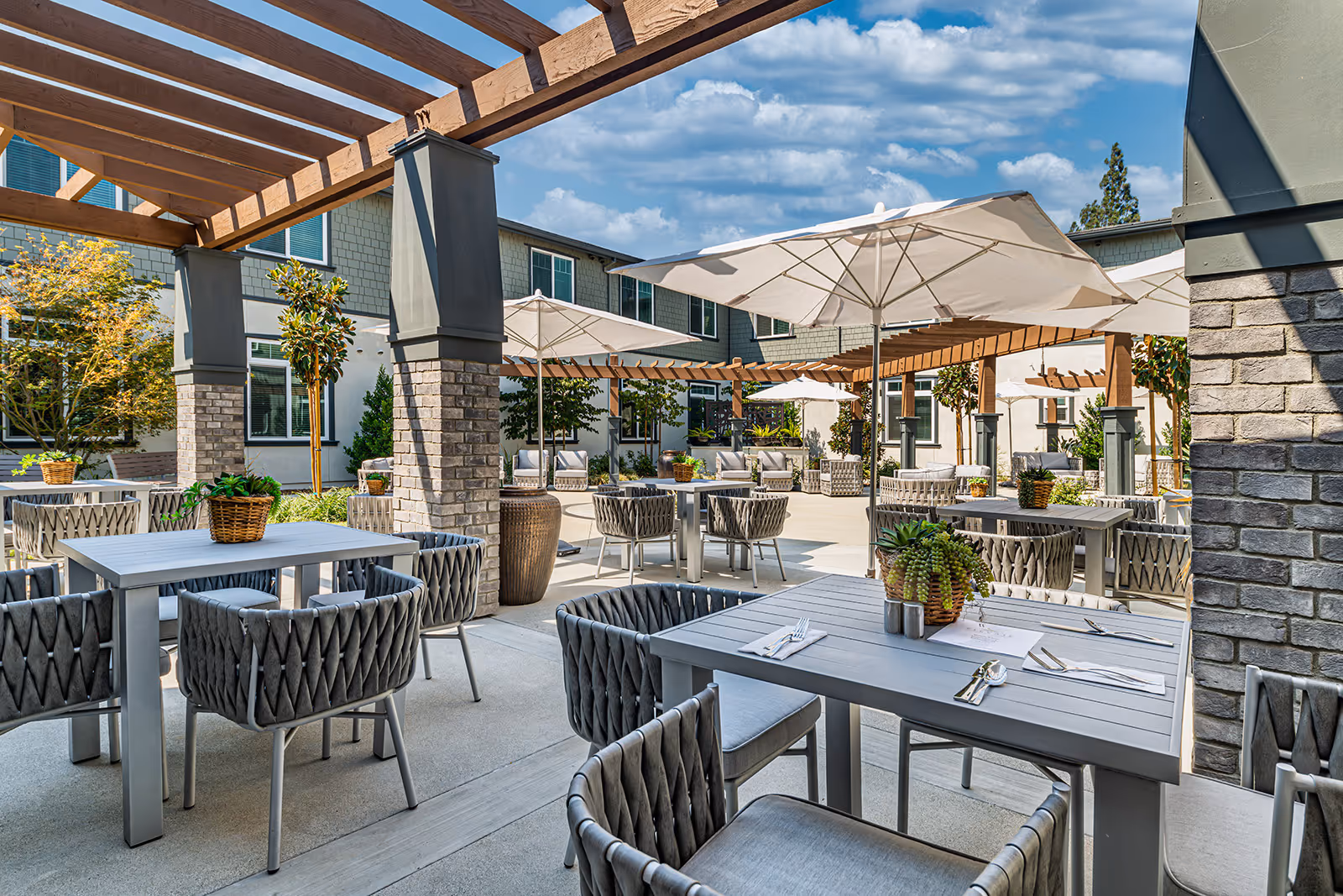 Outdoor patio area at Allara Senior Living with multiple tables and cushioned chairs under large white umbrellas and wooden pergolas, surrounded by greenery and a two-story building in the background under a partly cloudy sky.