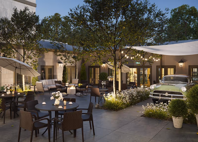 Outdoor courtyard area at Fields Senior Living at Smokey Point Assisted Living & Memory Care Community featuring multiple round tables with chairs, some with umbrellas, a seating area with sofas and armchairs, trees, decorative plants, string lights, and a vintage green car parked near the building entrance.
