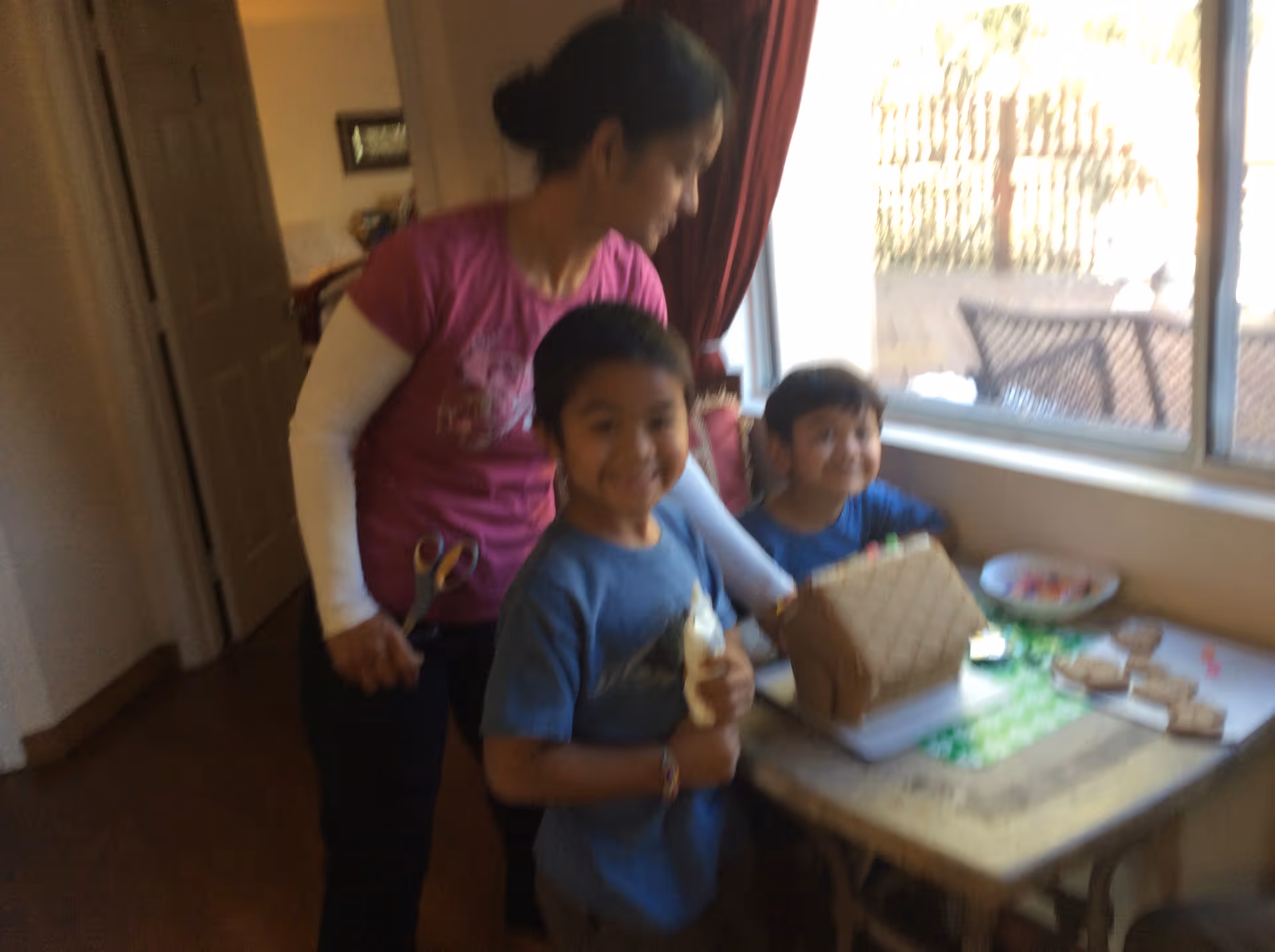 A woman and two children are gathered around a small table near a window. The children are smiling and appear to be decorating a gingerbread house. The woman is standing behind them, holding a pair of scissors.