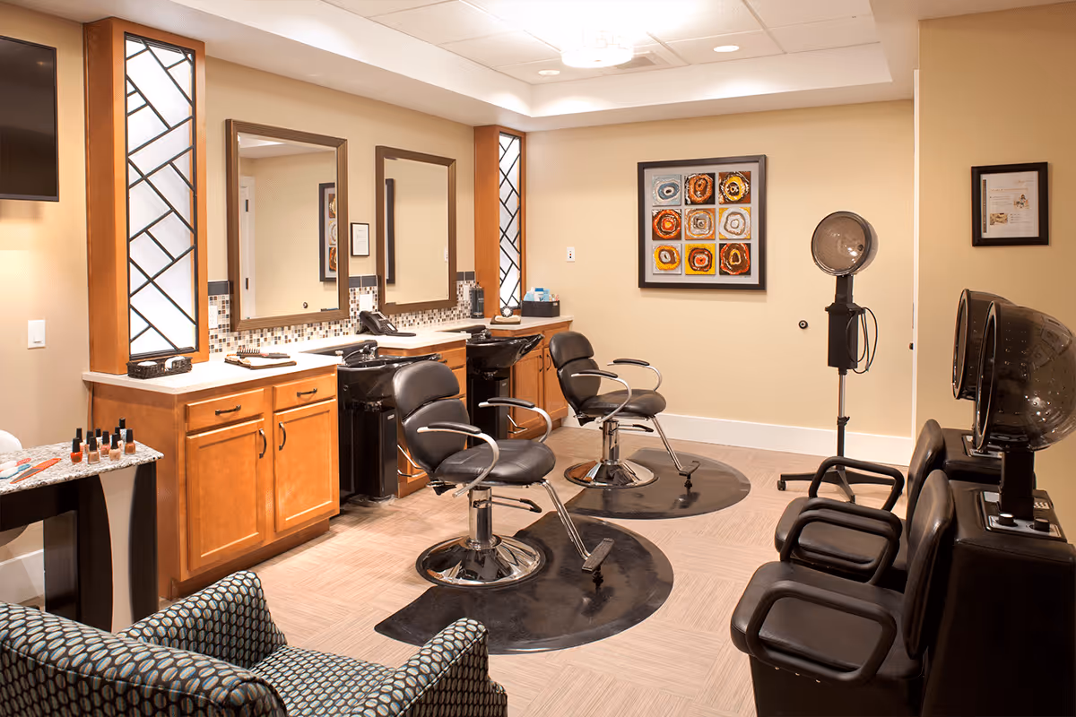 A well-lit salon room with two black salon chairs in front of sinks and mirrors, a patterned armchair, hair dryers, and a small table with nail polish bottles. The walls are beige with decorative framed artwork and mirrors.