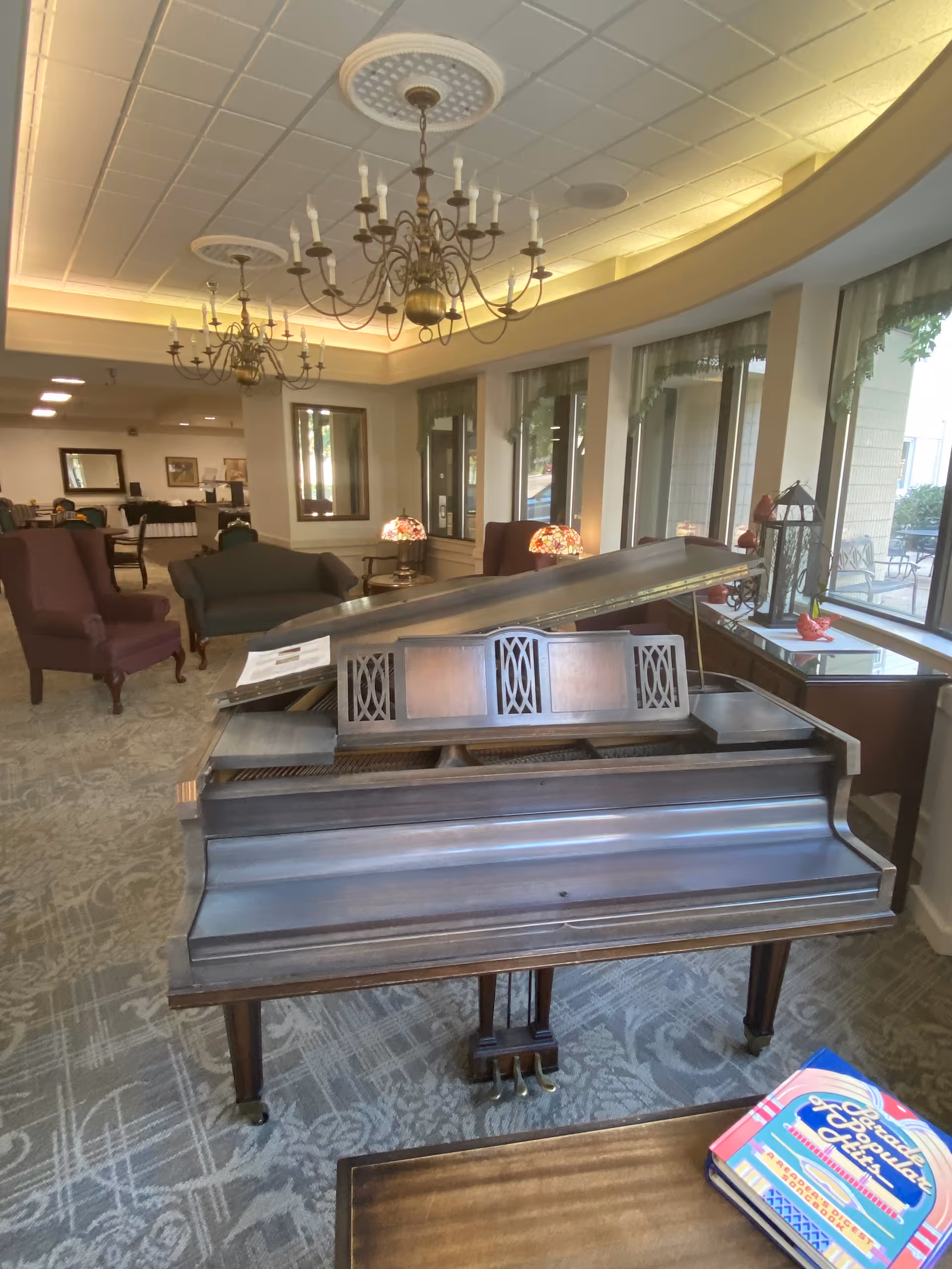Interior view of a senior living facility lounge area featuring a grand piano in the foreground, upholstered chairs and sofas, chandeliers hanging from the ceiling, large windows with sheer curtains, and decorative lamps on side tables.