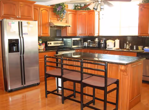A kitchen with wooden cabinets, a stainless steel refrigerator, an electric stove, a microwave, and various small kitchen appliances on the countertop. There is a kitchen island with a dark countertop and three black metal bar stools with brown cushions. The floor is wooden, and there is a window letting in natural light.