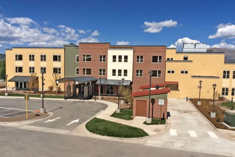 Exterior view of a multi-story senior living facility building with a combination of brick and yellow siding, a covered entrance, street lamps, a parking area, and a clear blue sky with some clouds.