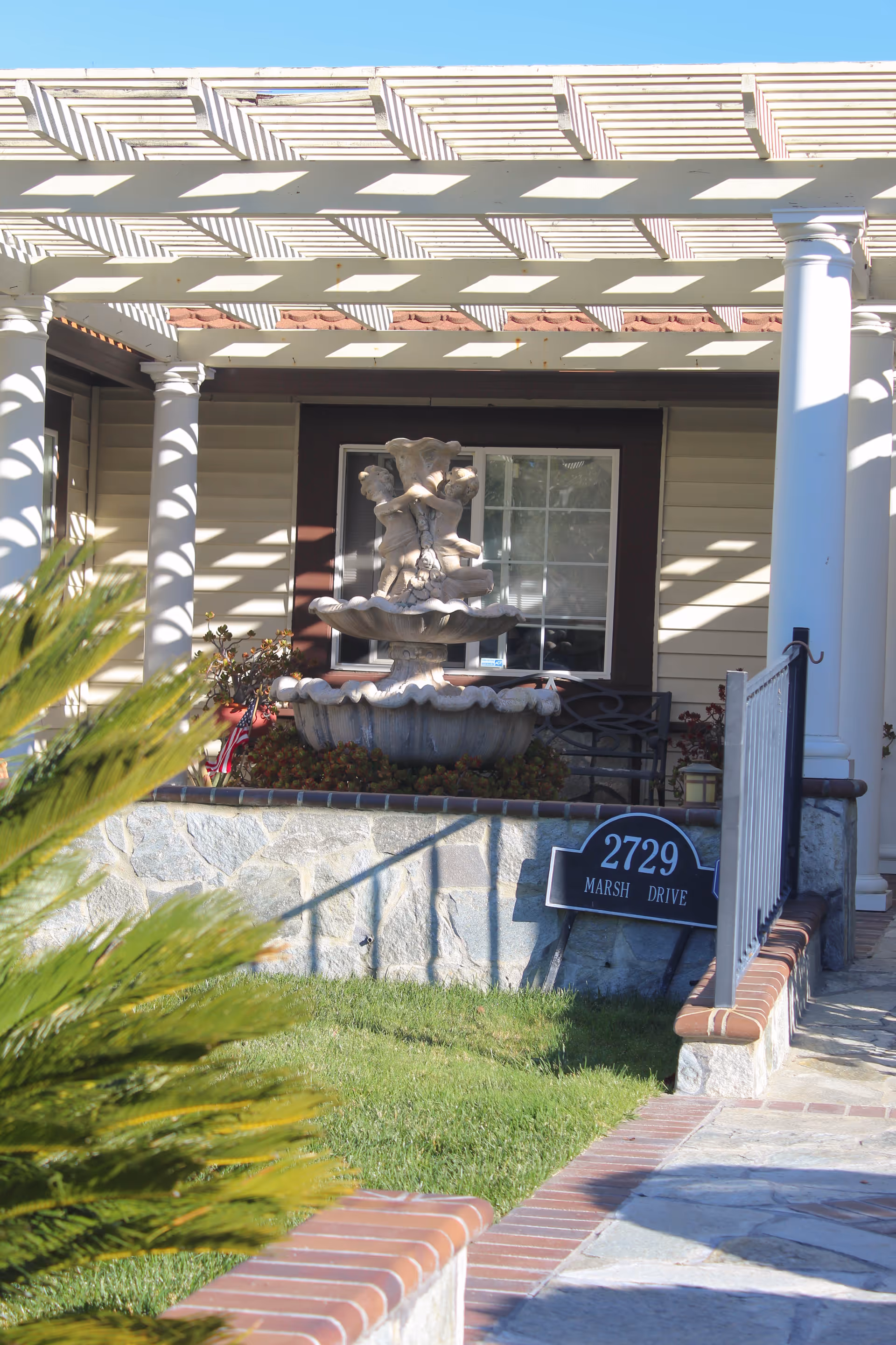 Front porch area of a building with a decorative two-tiered fountain featuring cherub statues, a window with white trim, white columns supporting a pergola casting striped shadows, a stone wall with a sign reading '2729 Marsh Drive', and a green lawn with some plants in the foreground.