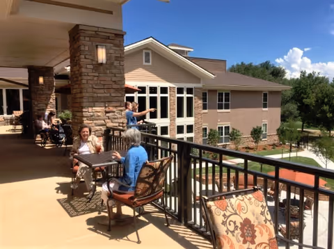 Two elderly women sitting and talking at a table on a spacious outdoor balcony of a senior living facility. The balcony has stone pillars, metal railings, and several chairs with patterned cushions. In the background, there is a multi-story building with large windows and a landscaped garden area under a clear blue sky.