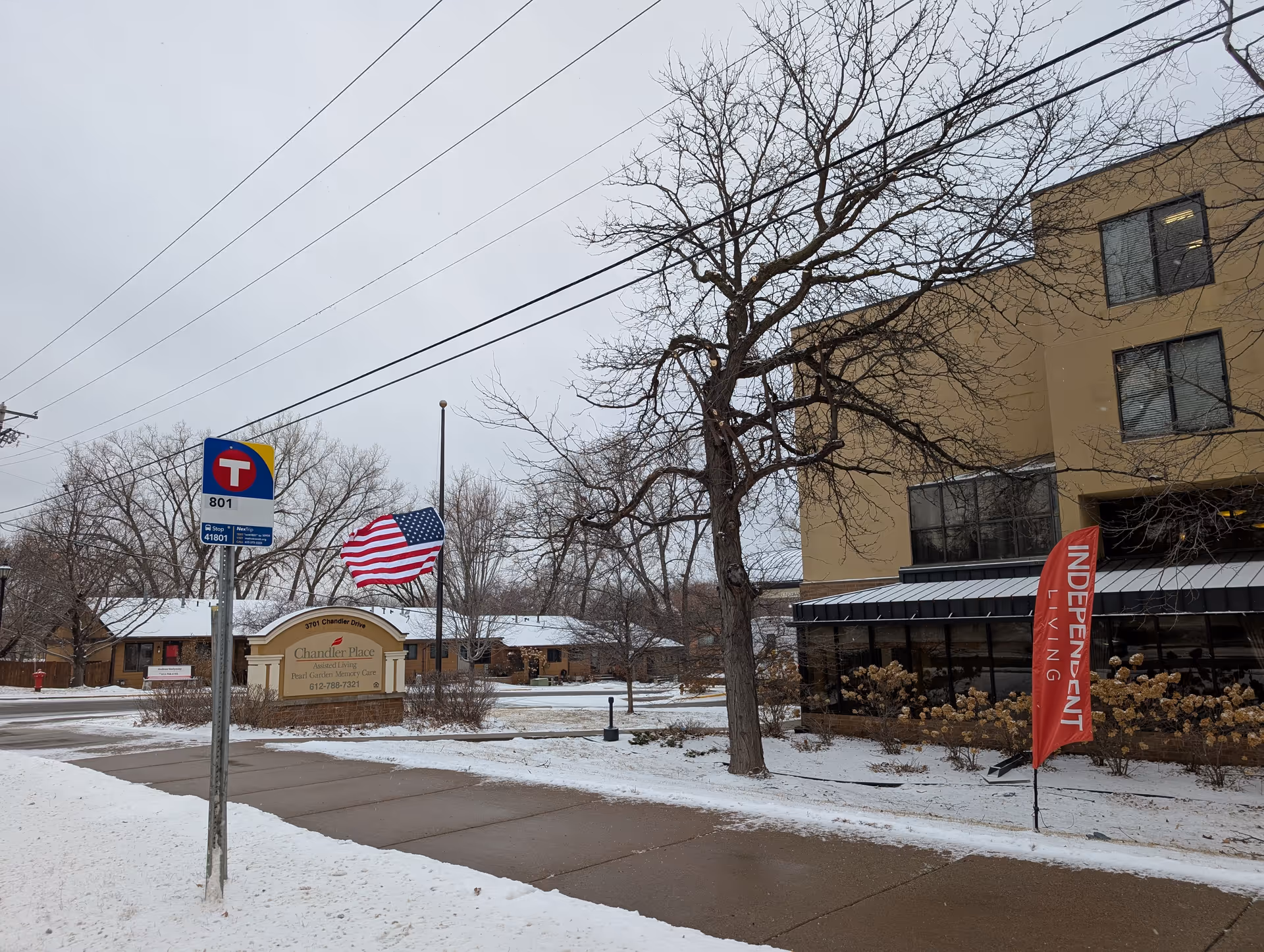 Exterior view of Chandler Place senior living facility during winter with snow on the ground. There is a sidewalk, a bus stop sign, an American flag on a flagpole, and a red flag that reads 'INDEPENDENT LIVING'. The building is tan with several windows and leafless trees around.