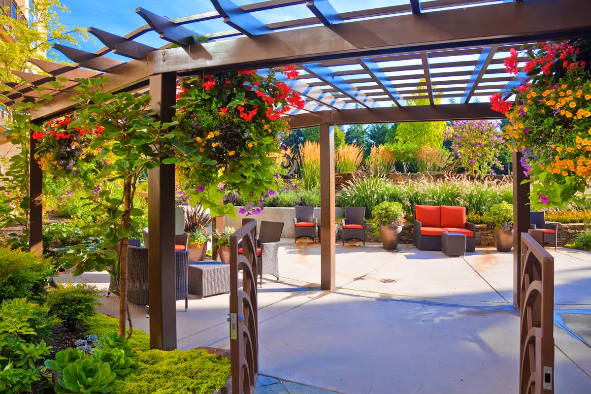 Outdoor patio area with a wooden pergola adorned with hanging flower baskets. The space includes wicker chairs and sofas with red cushions, surrounded by lush greenery and colorful plants under a clear blue sky.